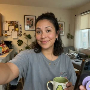Mujer sonriente con cabello oscuro tomando selfie en ambiente hogareño, sosteniendo taza y teléfono