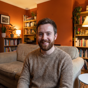 Hombre sonriente con barba sentado en sofá beige en living acogedor con paredes color terracota, estanterías de madera con libros, plantas decorativas y iluminación cálida que transmite tranquilidad y bienestar hogareño